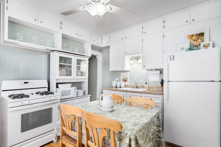 A Small, Old White Kitchen With White Whirlpool Appliances, A Ceiling Fan Hanging From The Ceiling, And A Small Dining Room Table With Four Wooden Chairs.