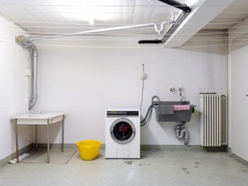 Shared Apartment Building Laundry Room With Washing Machine In Used Wash Sink And Piping Wide Angle View