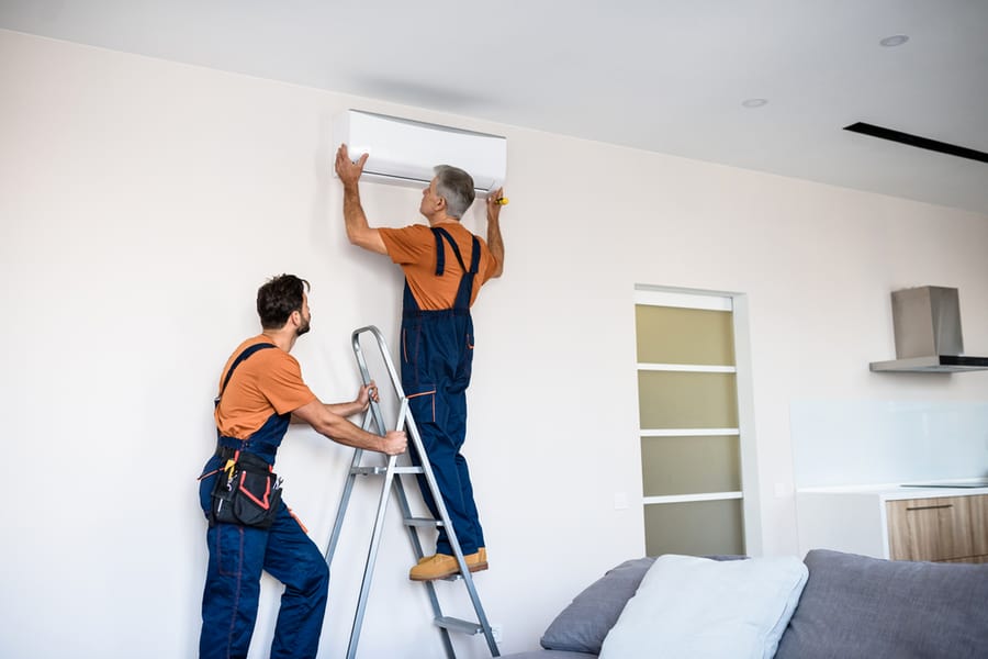 Two Workers In Uniform, Air Conditioning Masters Using Ladder While Installing A New Air Conditioner