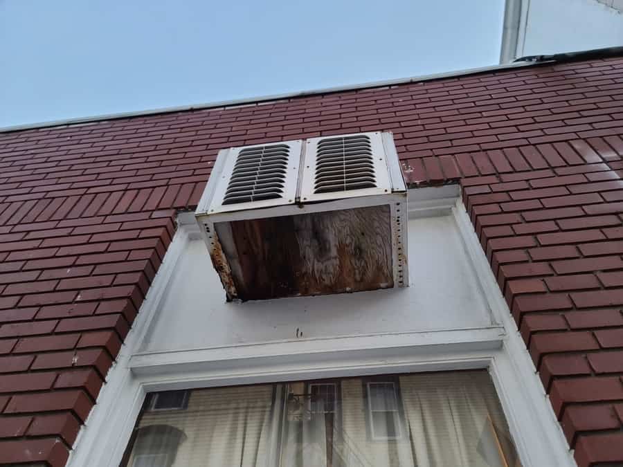 An Air Conditioner Sticking Out The Top Of A Winder With A Rotting Board Wedged Under It