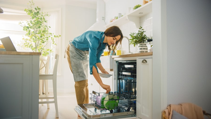 Beautiful Female Is Loading Dirty Plates Into A Dishwasher Machine In A Bright Sunny Kitchen