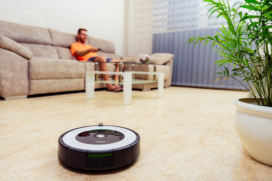 Man Sitting On A Sofa Watching Tv While The Irobot Roomba Cleans The Living Room Floor