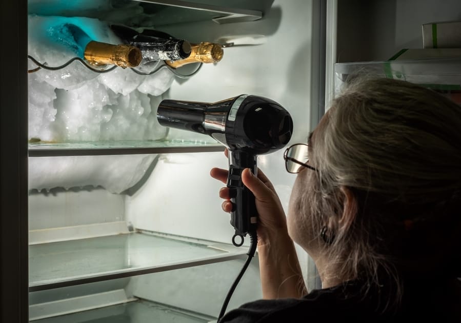 Woman Defrosting The Inside Of A Fridge Where There Are Some Stuck Bottles, With The Help Of A Hair Dryer.