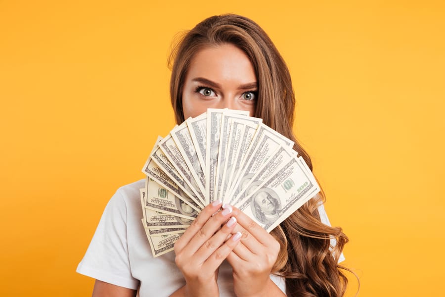 Close Up Portrait Of A Young Girl Covering Her Face With Money Banknotes Isolated Over Yellow Background.