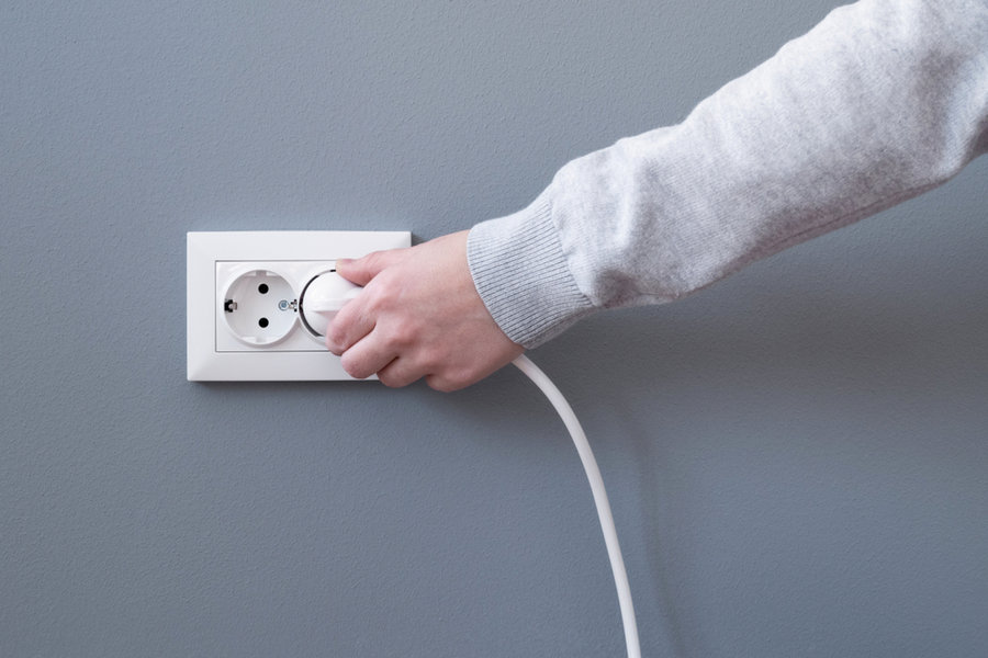 Hand Plugging In An Electric Cord Into A White Plastic Socket Or European Wall Outlet On Grey Plaster Wall. Closeup Of A Woman's Hand Inserting An Electrical Plug Into A Wall Socket. Daylight.