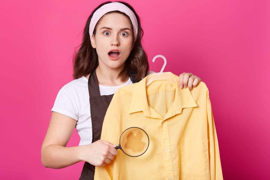 Image Of Shocked Female Wears White T Shirt, Brown Apron And Hair Band, Holds Yellow Shirt And Magnifier In Hand, Looks At Camera With Astonishment, Posing With Opened Mouth Against Pink Wall.