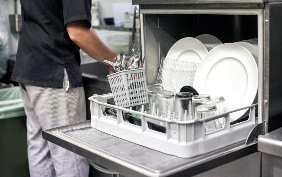 Kitchen Hand With An Open Dishwasher Filled With Clean White Plates In A Restaurant Kitchen In A Catering And Hygiene Concept