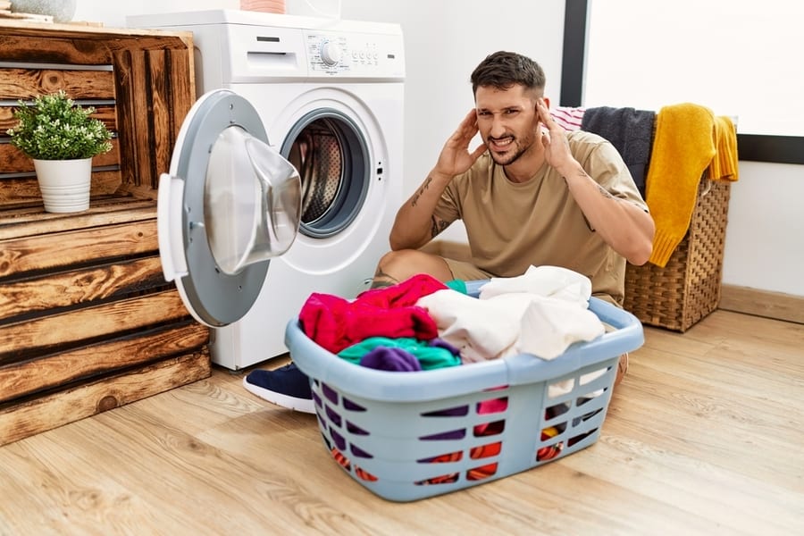 Man Putting Dirty Laundry Into Washing Machine Covering Ears With Fingers With Annoyed Expression