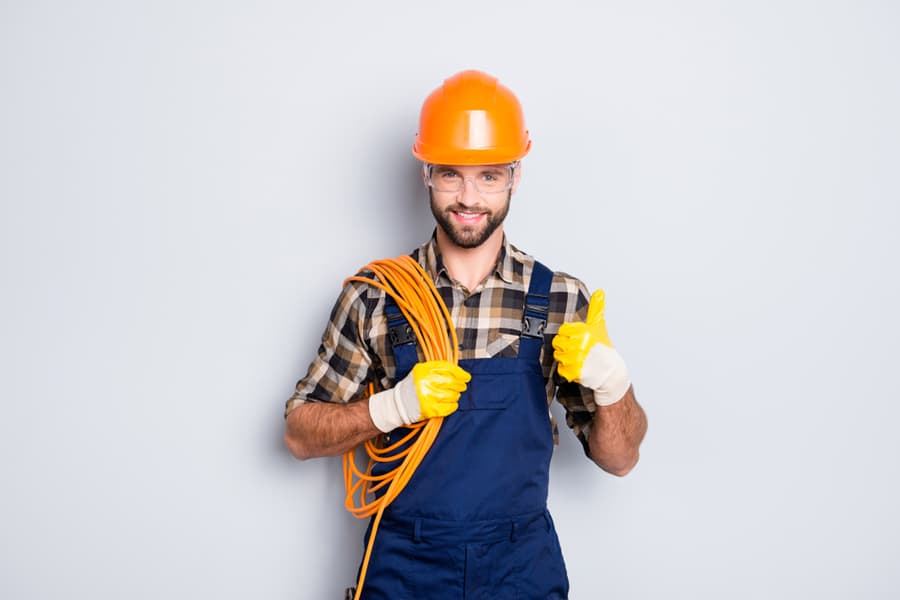 Portrait Of Handsome Joyful Electrician In Hardhat, Overall, Shirt With Bristle, Holding Rolled Wires On Shoulder, Showing Thumb Up Recommend Approve Sign Over Grey Background