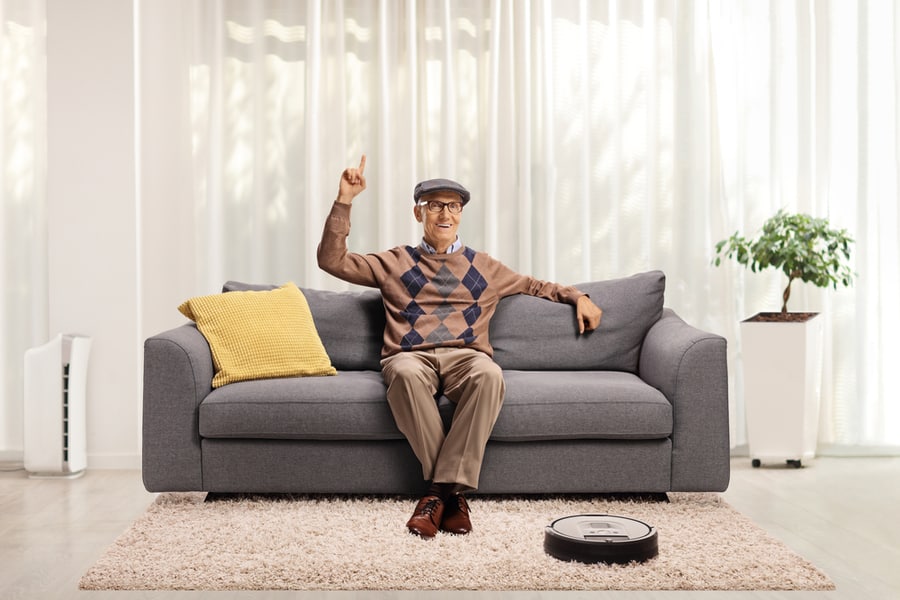 Smiling Elderly Man Sitting On A Sofa At Home, Pointing Up And A Robotic Vacuum Cleaner Dusting The Carpet