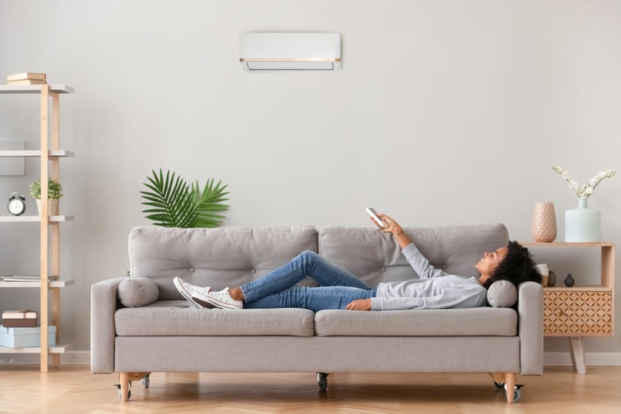 Woman In Casual Wear Lying On Sofa In Living Room At Modern Home Holding Air Conditioner Remote