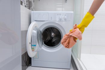 Yellow Rubber Gloved Hand Holding Dirty Pink Towel In Front Of Small Front Loading Washing Machine