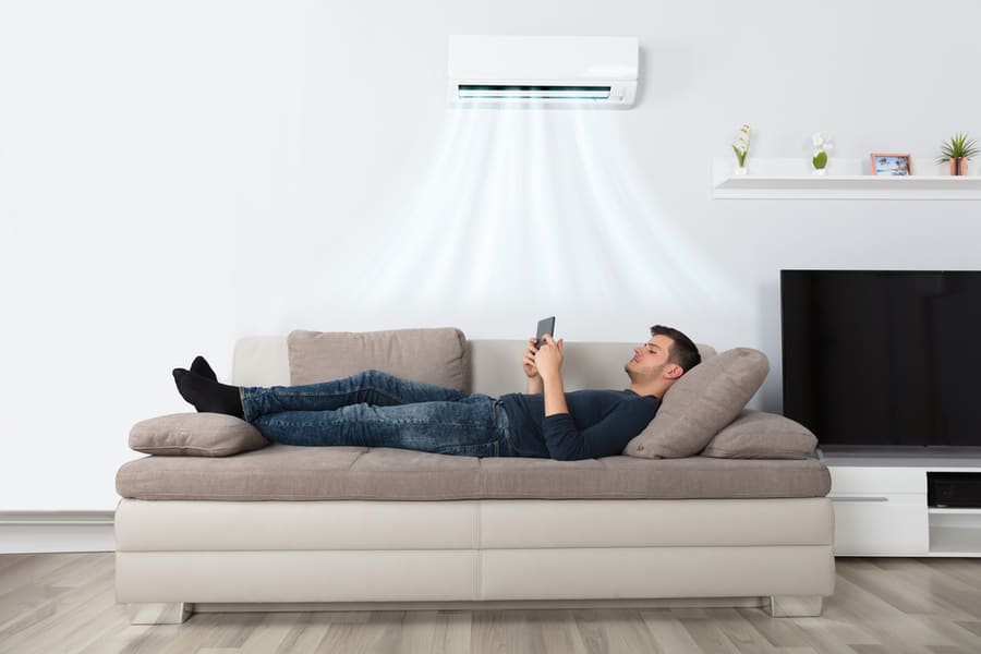 Young Man Lying Under Air Conditioner On Couch Using Tablet At Home