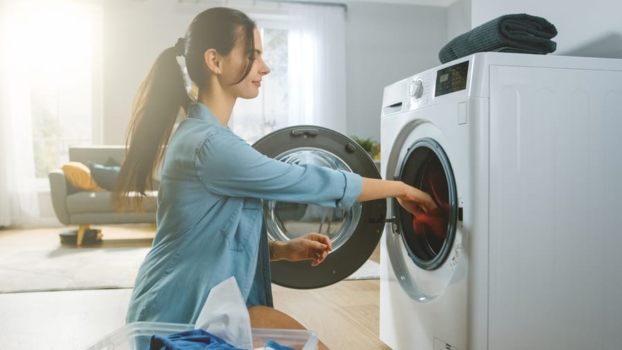 Young Woman Sits In Front Of A Washing Machine In Homely Jeans Clothes