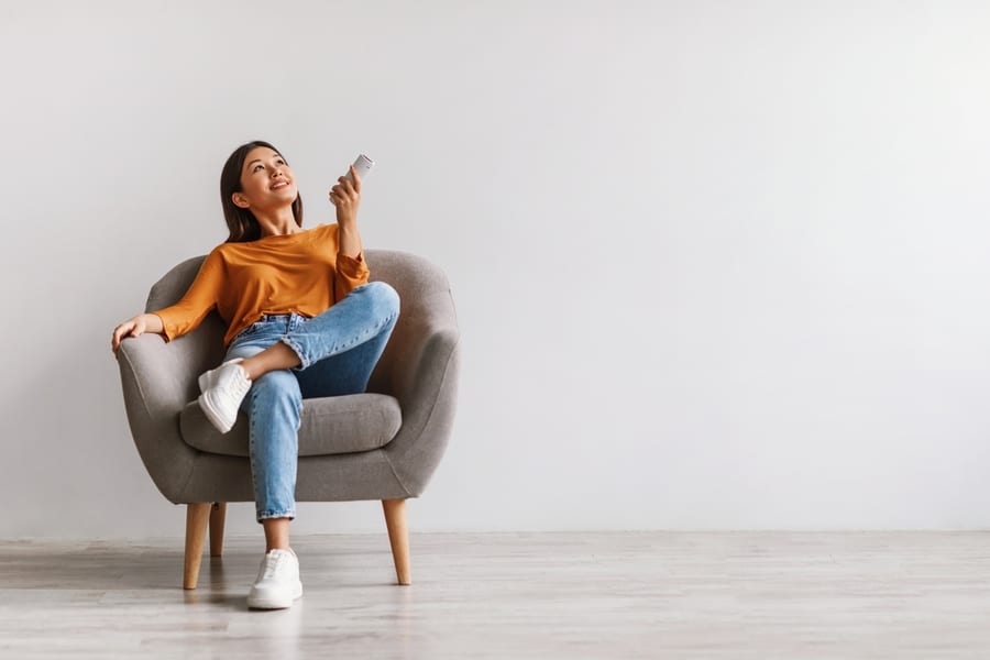 Happy Young Woman With Remote Relaxing Under Air Conditioner, Sitting In Armchair