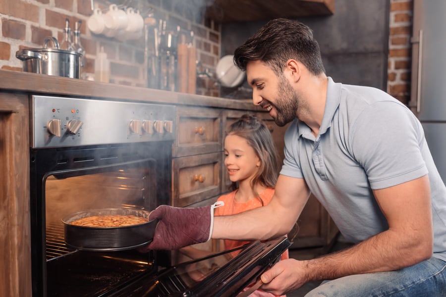 Side View Of Smiling Father And Daughter Taking Cake From Oven