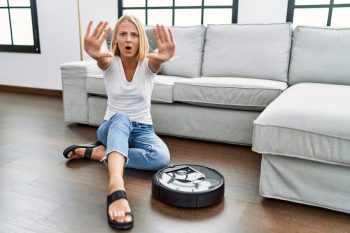 Woman Sitting At Home By Vacuum Robot Doing Stop Gesture With Hands Palms