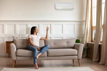 Young Asian Woman Switching On Air Conditioner While Resting Seated On Couch
