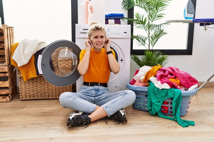 Young Blonde Woman Doing Laundry Sitting By Washing Machine Covering Ears With Fingers With Annoyed Expression