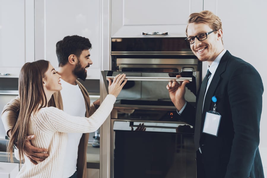 Young Couple Standing In A New Kitchen With A Salesman Inspecting The Oven